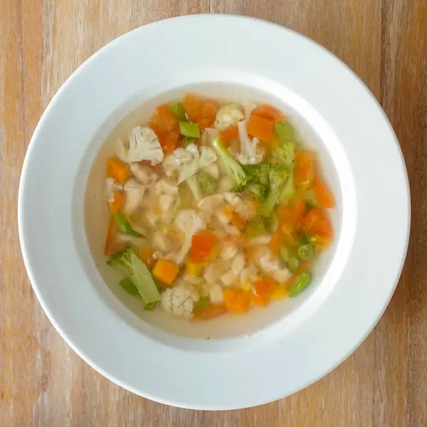 Temiz sebze çorbasıyla dolu beyaz bir seramik kase ahşap bir yüzeye oturur. Çorbada doğranmış portakal havuçları, yeşil brokoli çiçekleri ve beyaz karnabahar parçaları yer alıyor. Genel sunum basit ve sağlıklı bir yemek öneriyor.