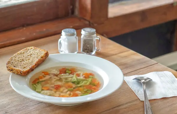 Brokoli, havuç ve dolma biber içeren temiz sebze çorbasıyla dolu beyaz tabak ahşap bir masada oturur. Tabağın kenarında bir dilim çoklu tahıllı ekmek duruyor. Yakınlarda tuzluk ve biberlik ve kaşık beyaz bir peçeteye konur..