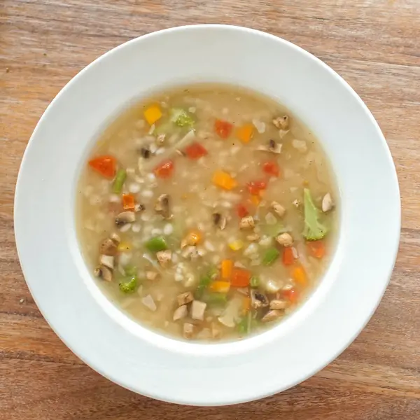 Ahşap bir yüzeye sebze çorbası ve arpa dolu beyaz bir kase yerleştirilir. Çorba hafif ve temiz görünüyor, arpa taneleri etrafa dağıtılmış. Sunum basit, yemeğin kırsal ve sağlıklı doğasını vurguluyor.