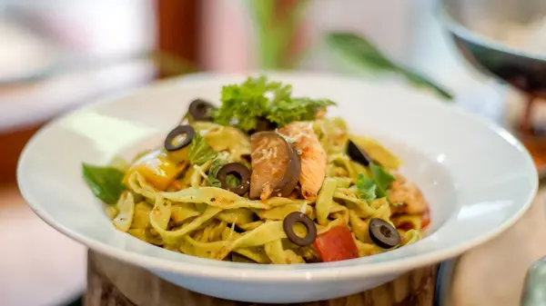 Beyaz bir tabakta servis edilen renkli makarna, üzerinde siyah zeytin dilimleri, sarı dolma biber ve taze maydanoz bulunan tagliatelle..