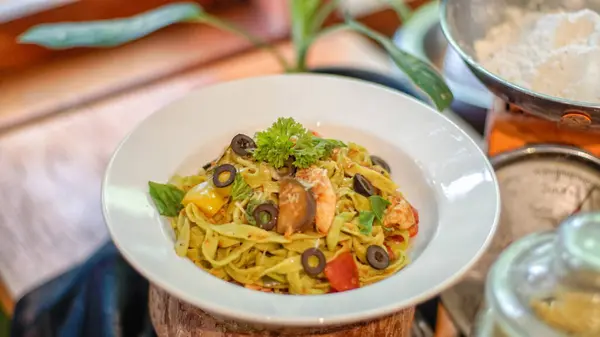 Beyaz bir tabakta servis edilen renkli makarna, üzerinde siyah zeytin dilimleri, sarı dolma biber ve taze maydanoz bulunan tagliatelle..