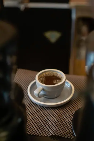Koyu sıcak bir içecekle dolu beyaz bir seramik fincan, muhtemelen kahve, uyumlu bir çay tabağının üzerinde duruyor. Tezgahın üzerindeki desenli, kareli bir paspasın üzerine yerleştirilmiş.