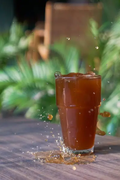 Bir bardak koyu kahverengi sıvı, muhtemelen çay ya da kola, ahşap bir masada oturur. Sıvı fışkırıyor, havada damlacıklar ve masada bir sızıntı yaratıyor. Arkaplan özellikleri bulanık yeşil yeşillik, açık hava ayarlarını gösterir.