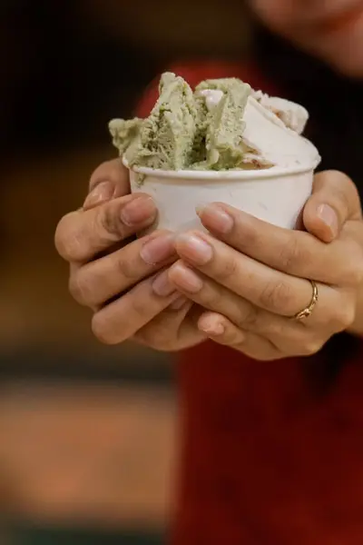 İki kepçeli bir fincan dondurma, biri yeşil ve biri beyaz. Arka plan hafifçe bulanık, altıgen ışıklar bir bokeh etkisi yaratıyor, dondurma ve ellere odaklanmayı arttırıyor..