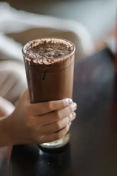 Bir el soğuk çikolata içeceği ile dolu uzun bir bardağı tutar. İçecekler köpüklü görünüyor, yüzeyinde de çikolata lekeleri var. Arkaplan bulanık