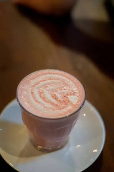 Bir bardak kırmızı kadife latte ahşap bir masanın üzerinde beyaz bir fincan tabağı üzerine konur. Lattelerin yüzeyi, kırmızı ve beyaz kıvrımlı karmaşık köpük sanatı sergiliyor. Görsel olarak çekici bir desen oluşturuyor..
