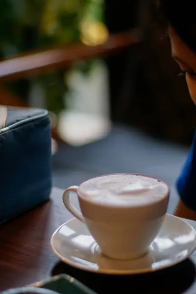 Köpüklü bir içecekle dolu beyaz bir seramik fincan, koyu ahşap bir masanın üzerinde, uyumlu bir fincan üzerinde duruyor.