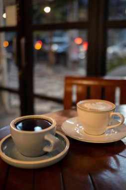 Sıcak bir kafede, ahşap bir masada iki fincan oturuyor. Sol fincan açık mavi ve koyu kahve içerir, sağ fincan ise beyaz, köpüklü sütlü ve üstünde latte sanatıyla birlikte..