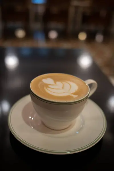 Beyaz bir seramik fincan ve çay tabağı parlak siyah bir masada bir latte tutuyor. Latte, stilize edilmiş bir kalp ve yaprak desenini andıran karmaşık köpük sanatını içeriyor. Ayarlar, bulanık arkaplanı olan sıcak ve rahat bir ortam izlenimi veriyor. 