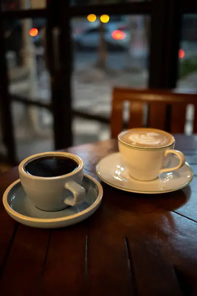 Sıcak bir kafede, ahşap bir masada iki fincan oturuyor. Sol fincan açık mavi ve koyu kahve içerir, sağ fincan ise beyaz, köpüklü sütlü ve üstünde latte sanatıyla birlikte..
