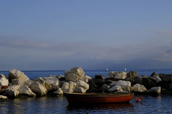 Small red wooden fishing boat. The boat was placed in the small port of Naples bordered by cliffs. In the background the Lattari Mountains covered by clouds. Photo taken during a winter sunset.