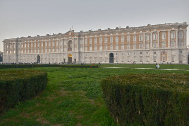 The facade of the Royal Palace of Caserta portrayed from the front garden in Piazza Carlo di Borbone. The hedges of the green area of the square can be seen in the foreground. Photo taken during the blue hour.