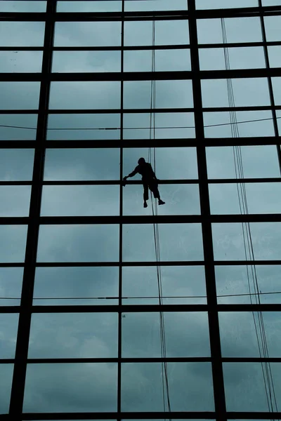 A silhouette of a worker cleaning windows hangs from a rope in the airport building.