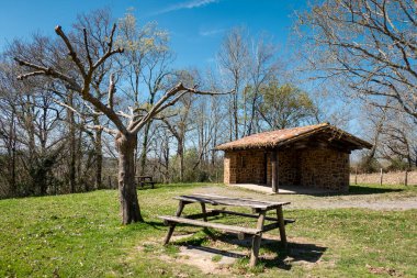 Park next to the Chapel de San Salvador in Jatxou with a table and wooden benches in the middle of the grass, with a small garden house with a bathroom in the background 