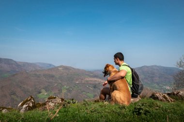 Young tourist sitting relaxing with his dog and enjoying the mountains and villages in the background from the Jara mountain in Iroulguy in the Basque Country clipart