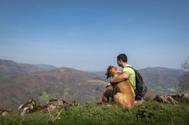 Young tourist sitting relaxing with his dog and enjoying the mountains and villages in the background from the Jara mountain in Iroulguy in the Basque Country clipart