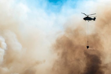 Helikopter çam ağacında yanan bir orman yangını üzerine su boşaltıyor. Büyük beyaz ve siyah bir duman bulutu bırakıyor.