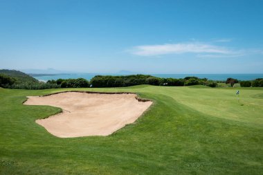 Güneşli bir günde arka planda ağaçlar ve deniz olan yeşil bir golf sahasının ortasındaki sığınaklar veya kum taneleri.