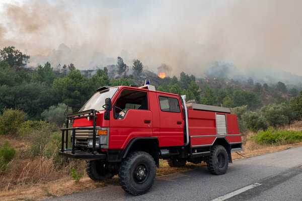 Fire engine next to the road fighting a forest fire with large flames that leave a large cloud of smoke in the air