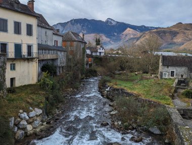 Soğuk ve bulutlu bir günde arka planda büyük dağlarla küçük bir köyün ortasında akan akarsu.