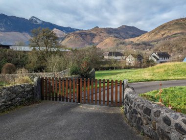 Yolun ortasında kırsal taştan bir duvarı olan ahşap bir kapı, soğuk bir günde arka planda bazı evler ve dağlar ve çok bulutlu bir gökyüzü.