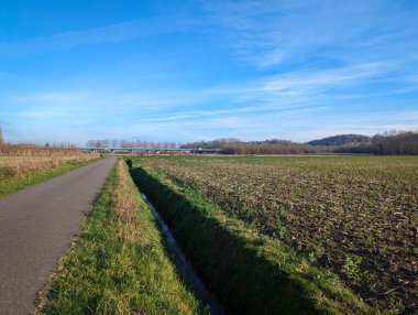 Güneşli bir günde, tarımın ortasında bir su kemeriyle çevrili küçük asfalt yol.