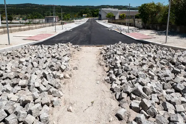 stock image Construction site with a road under restoration with new asphalt flooring with a continuation of parallel floors made of small granite cubes in the middle of a village