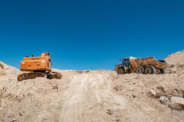 Çapa ve çöp kamyonu engebeli bir arazinin ortasında, arka planda mavi gökyüzü olan bir taş ocağında çalışıyor.
