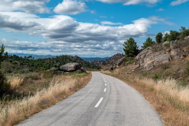 Dağlar, kayalar ve ormanlar arasında uzanan yol mavi gökyüzünün altında bazı bulutlarla benekli.