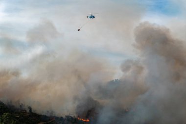 Helikopter havada duman bulutları bırakan bir orman yangınıyla mücadele ediyor.