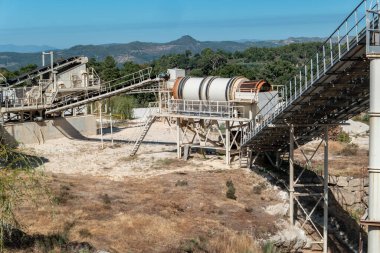 Arkasında orman ve dağ manzarası olan bir taş ocağında malzeme işleme makineleri olan bir fabrika.