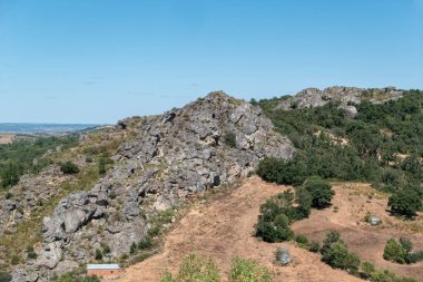 Mavi gökyüzünün altında büyük kaya oluşumları, ağaçlar ve kırsal alanlara sahip doğal manzara.