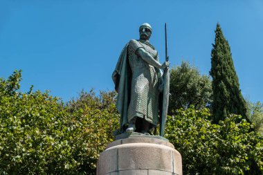 Portekiz 'in ilk kralı D. Afonso Henriques' in bronz heykeli. Mavi gökyüzünün altında, yoğun yeşil bitki örtüsüyle çerçevelenmiş zırh ve kılıç giyiyor.