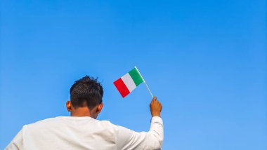 Boy holding Italy flag against clear blue sky. Man hand waving Italian flag view from back, copy space for text