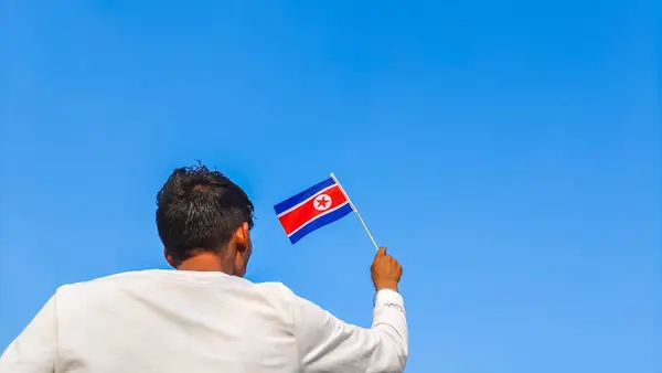 Boy holding North Korea flag against clear blue sky. Man hand waving North korean flag view from back, copy space for text