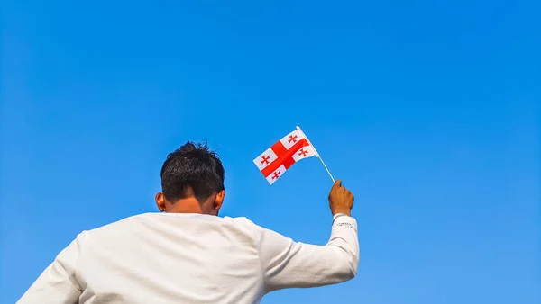 Boy holding Georgia flag against clear blue sky. Man hand waving Georgian flag view from back, copy space for text