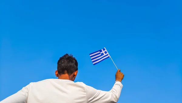 Boy holding Greece flag against clear blue sky. Man hand waving flag of Greece view from back, copy space for text