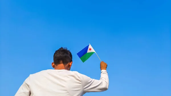 Boy holding Djibouti flag against clear blue sky. Man hand waving flag of Djibouti view from back, copy space for text