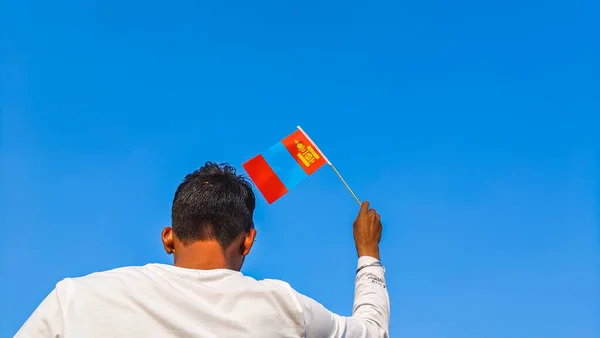 Boy holding Mongolia flag against clear blue sky. Man hand waving Mongolian flag view from back, copy space for text