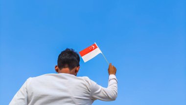 Boy holding Singapore flag against clear blue sky. Man hand waving flag of Singapore view from back, copy space for text