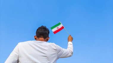 Boy holding Iran flag against clear blue sky. Man hand waving Iranian flag view from back, copy space for text