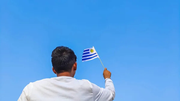 Boy holding Uruguay flag against clear blue sky. Man hand waving Uruguayan flag view from back, copy space for text