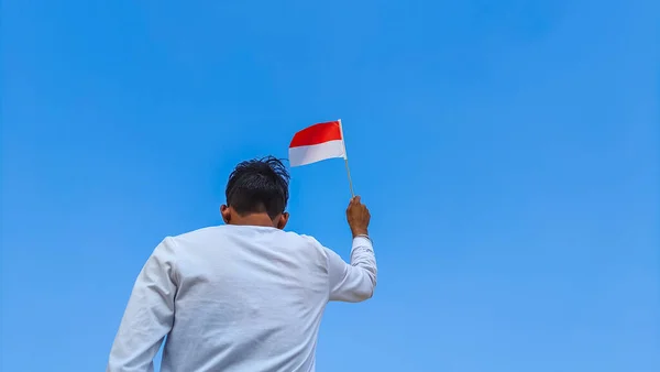 Boy holding Indonesia flag against clear blue sky. Man hand waving Indonesian flag view from back, copy space for text