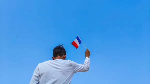 Boy holding France flag against clear blue sky. Man hand waving French flag view from back, copy space for text