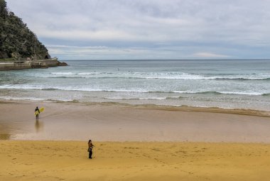 Sahilde yürüyen yalnız bir kadın ve denize giren sörfçü.