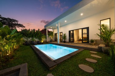 Bahçeli tropik villa manzarası, yüzme havuzu ve gün batımında açık oturma odası..