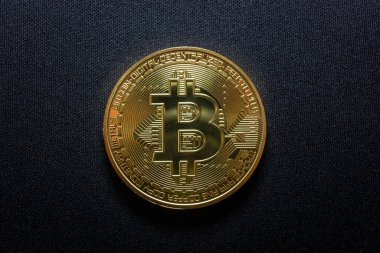 Koyu dokulu bir yüzey üzerinde duran fiziksel bir Bitcoin madeni parasının yakın plan çekimi. Altın sikke, merkezinde simgesel Bitcoin 