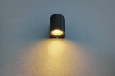 Modern, silindirik, koyu gri ya da siyah renkli bir lamba, bembeyaz ya da açık renkli, aşağı doğru parlayan bir duvarı aydınlatır..
