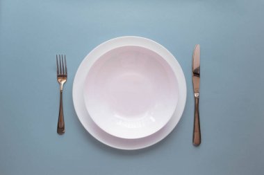 Açık mavi arka planda boş beyaz tabak, çatal ve bıçak. Üst görünüm, alanı kopyala.
