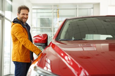 Erkek müşteri kırmızı arabanın yanında duruyor, kameraya bakıyor ve gülümsüyor. Popüler araba galerisinde satılık otomobili seçen adam. Sarı ceketli ve sakallı..